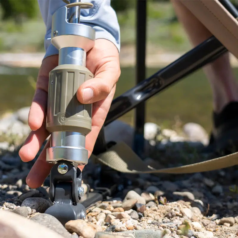 Hand adjusts the tan suspension leg of an Adjustable Rocker chair resting on a bed of rocks.