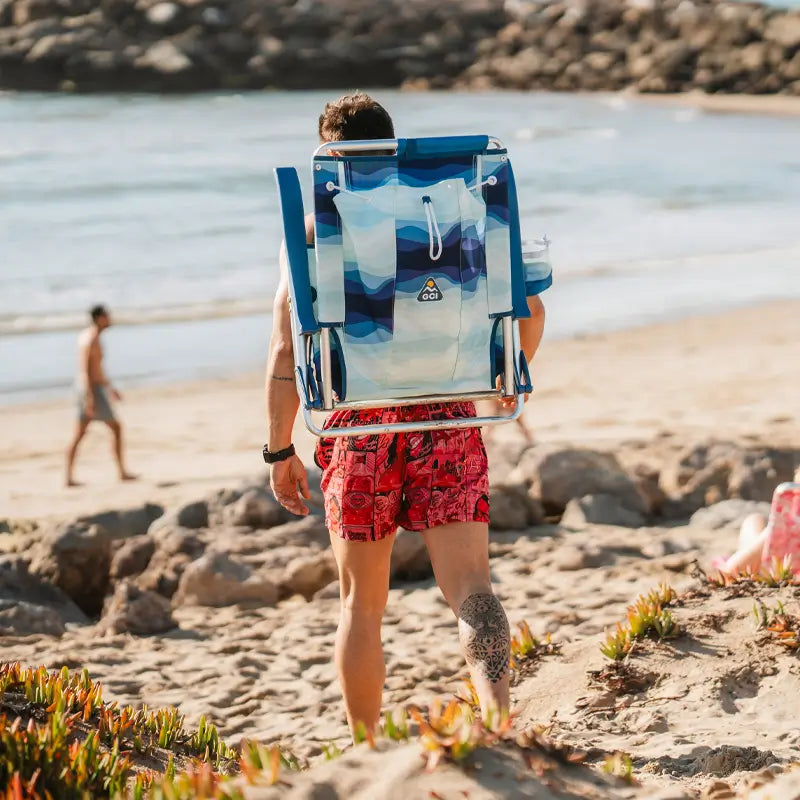 Man walking on the beach with a folded Wave variant Backpack Beach Chair on his back, showing blue gradient wave pattern.