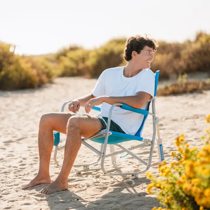 Man seated and smiling in a Saybrook Blue Beach Rocker on a sandy beach surrounded by coastal vegetation.