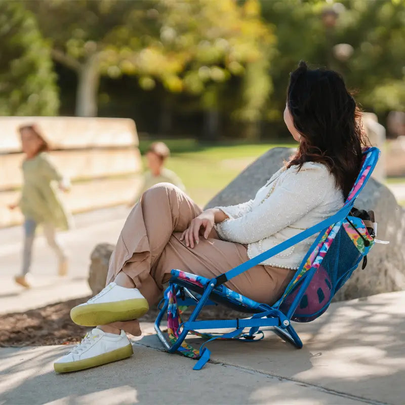 Woman relaxing in a tie dye Everywhere Chair 2 while kids play nearby in a park setting.