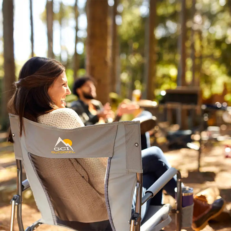 Smiling woman lounges in a pewter Firepit Rocker at a sunny forest picnic.