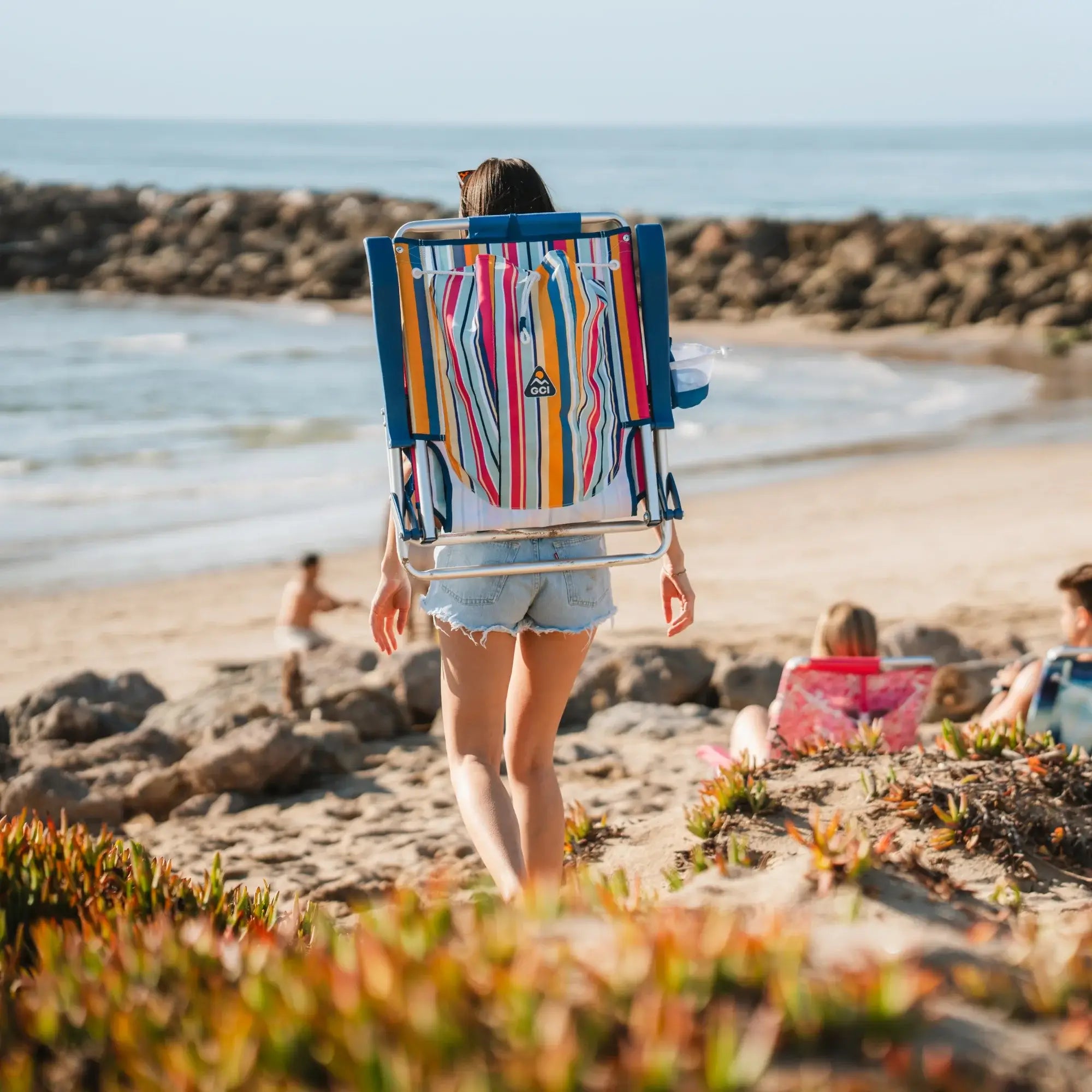 A woman walking down towards the ocean with the backpack beach chair attached on her back.