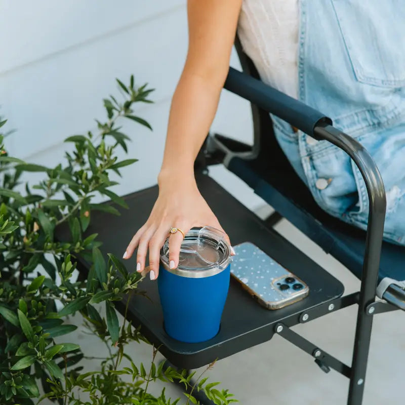 Close-up of a woman’s hand resting on the Slim-Fold Directors Chair side table with a blue tumbler and phone.
