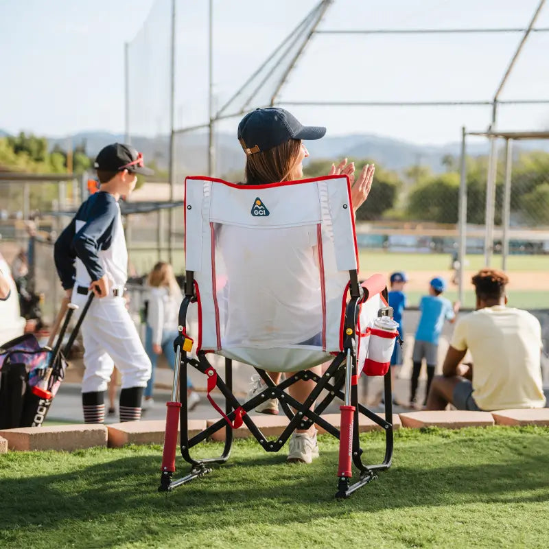 Person sitting on a folding chair at a baseball field with other people in the background.