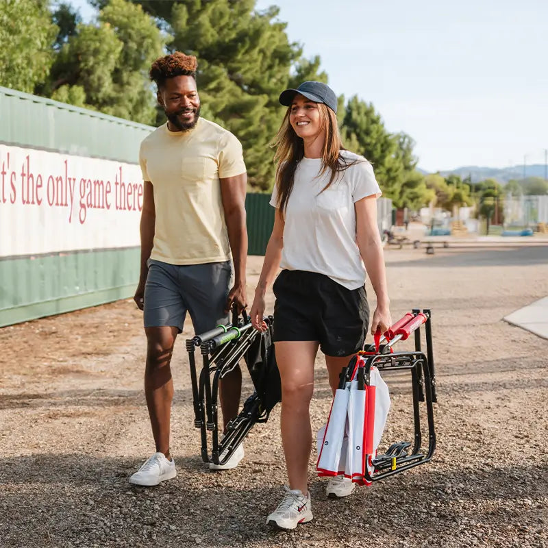Two people standing outdoors with sports equipment, holding a folding chair and a bag.