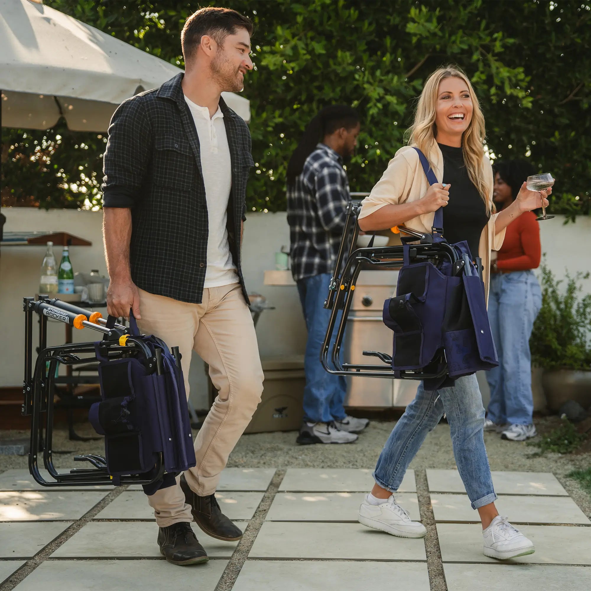 Two people laughing and walking while holding blue tailgater stowaway rockers. One person holding with a shoulder strap and another person using the carry handle.