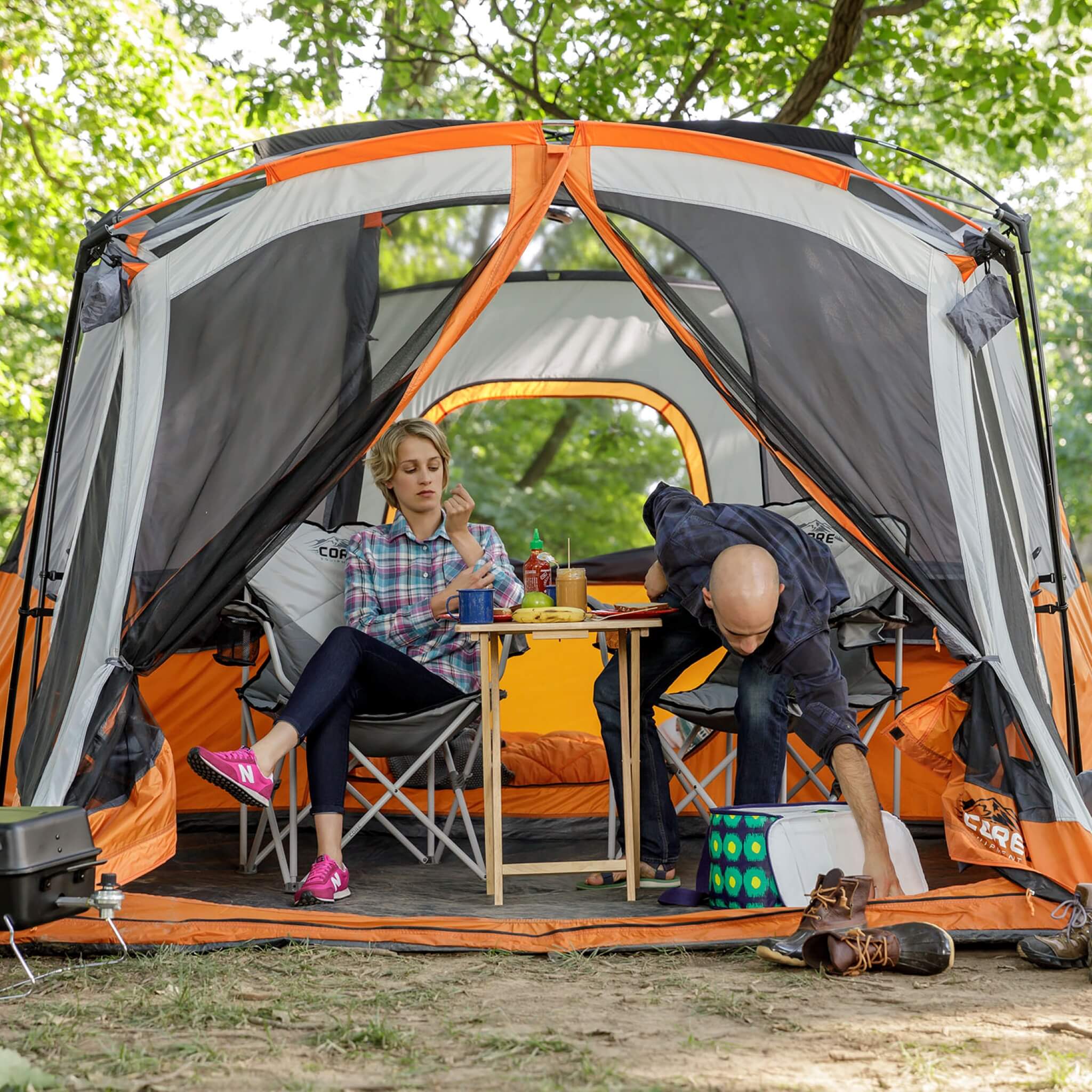 CORE 11 person straight wall cabin tent with screen room in orange and gray set up at a campsite with campers relaxing under inside the tent.