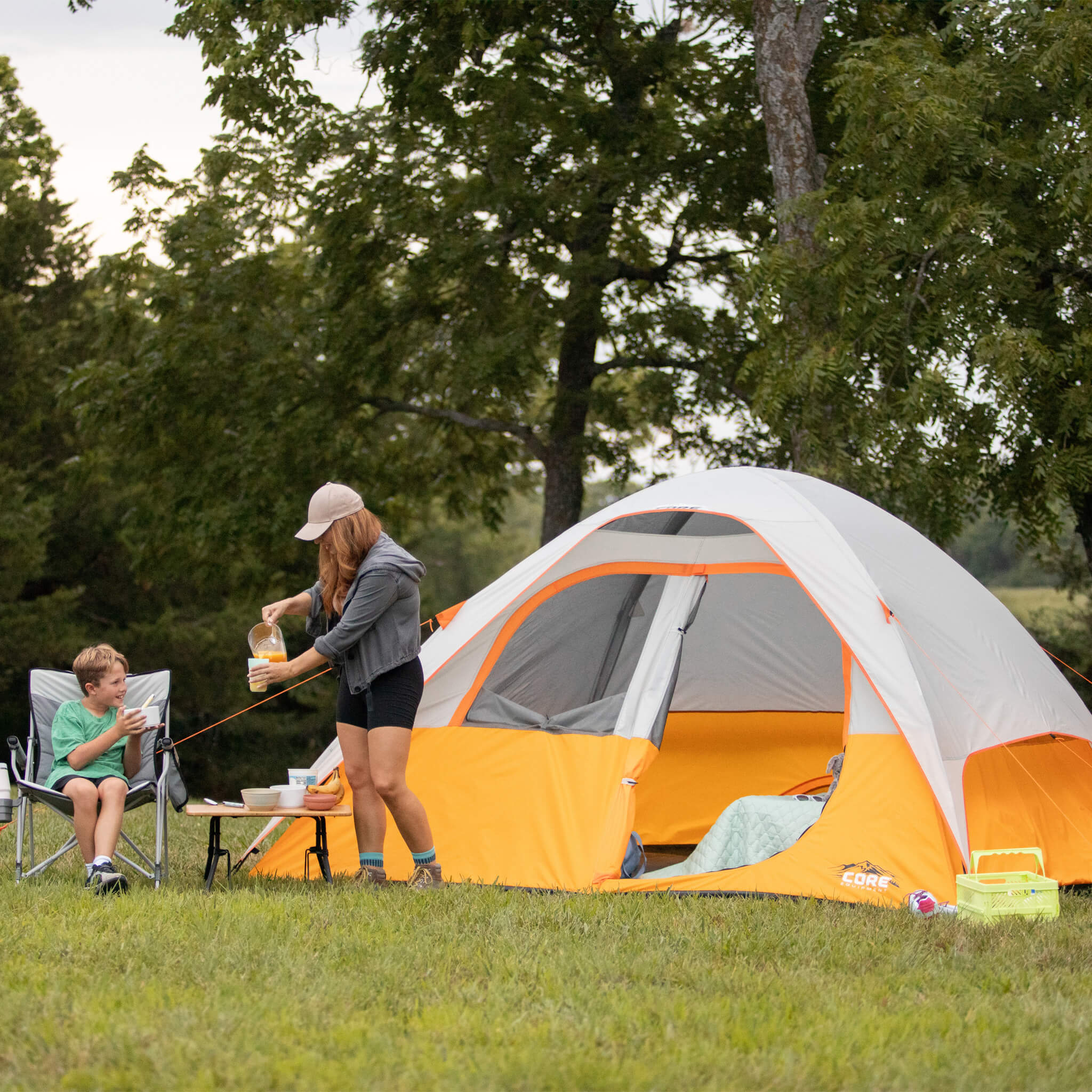 A mother and son camping with a CORE 6 person dome tent set up on a grassy campsite, showing campers enjoying outdoor activities.