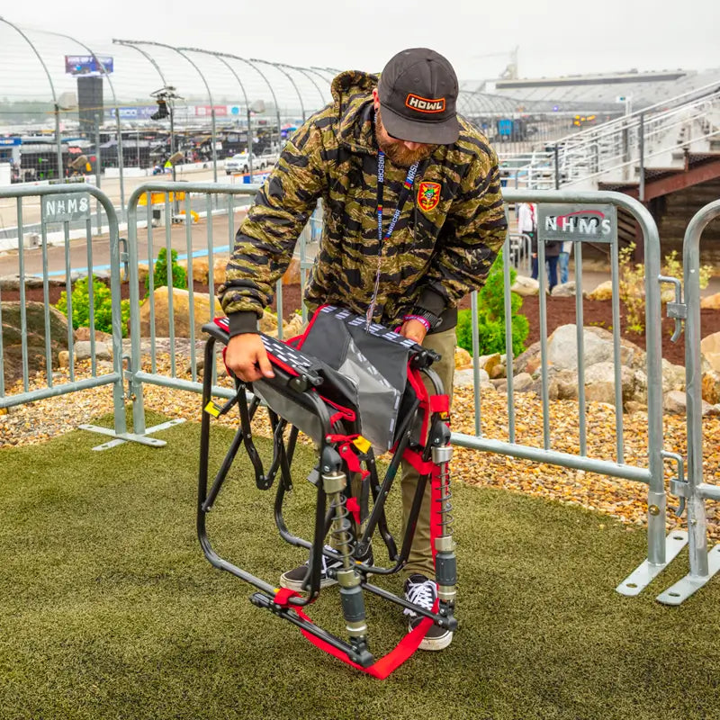 Man in a camo jacket folds up a red-trimmed Adjustable Rocker at a racetrack venue.