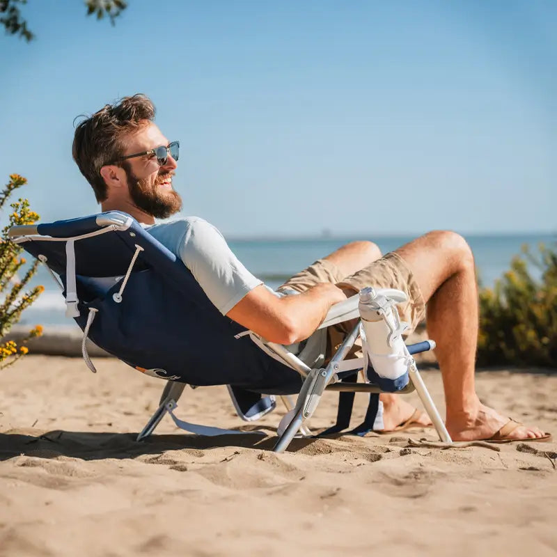 Man reclining in a Nautical Blue Backpack Beach Chair on the sand, smiling and enjoying a sunny beach day.