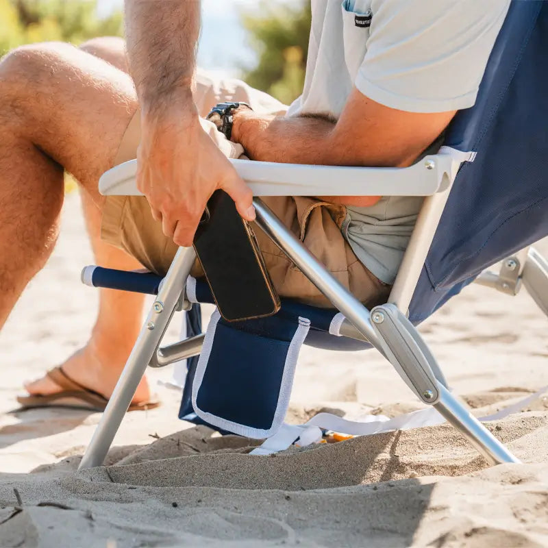 Man placing a phone into the side storage pocket of a Nautical Blue Backpack Beach Chair on the beach.