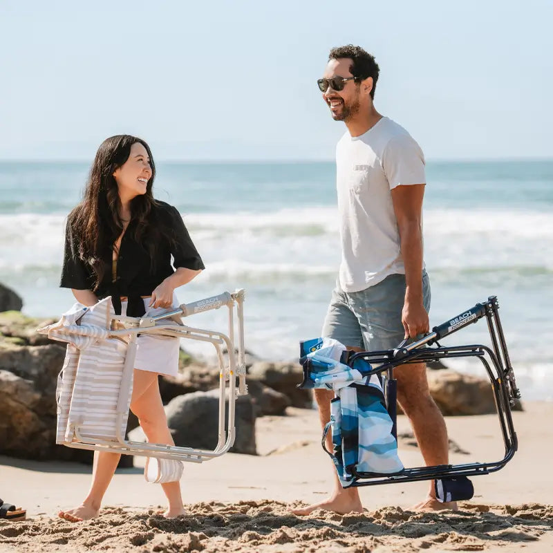 Couple walking on the beach carrying folded Blue Beach Wave and beige beach chairs with towels draped over them.