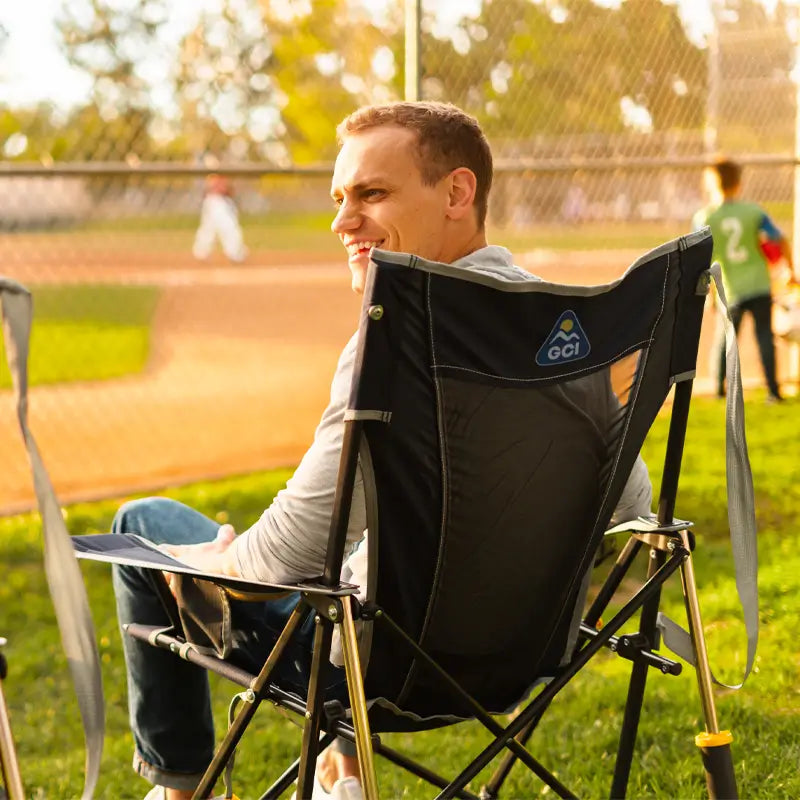 Man smiling in a GCI Comfort Pro Rocker Indigo chair while watching a baseball game at the park.