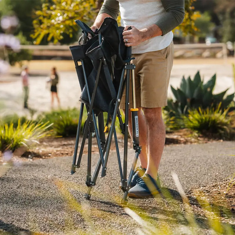 Man unfolding a Heathered Charcoal Comfort Pro Rocker XL chair on a sunny outdoor path.
