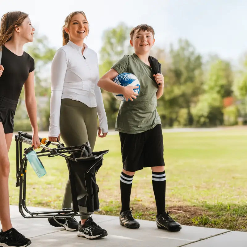 A family walks on a sidewalk at a park, carrying a folded black Freestyle Rocker and a soccer ball.