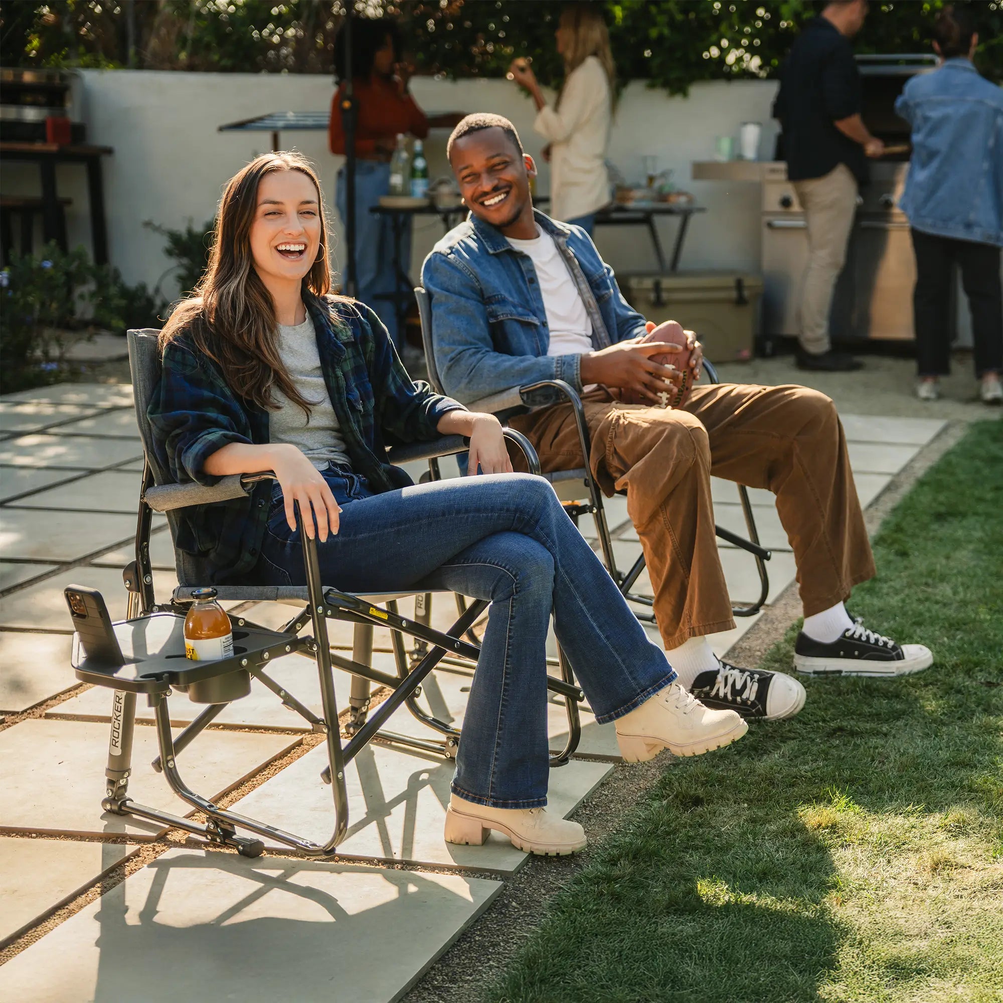 Two people smiling and laughing while sitting in freestyle rockers with side tables on a concrete patio. 