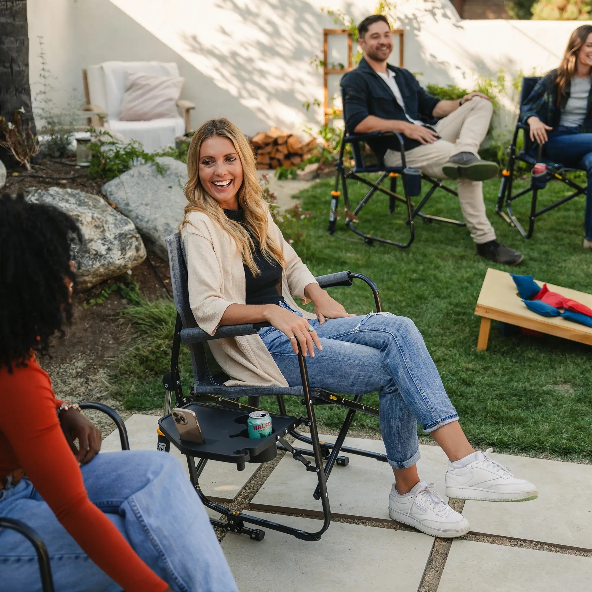 A woman smiling while conversing with another woman while they both site in freestyle rocker with side table chairs. 