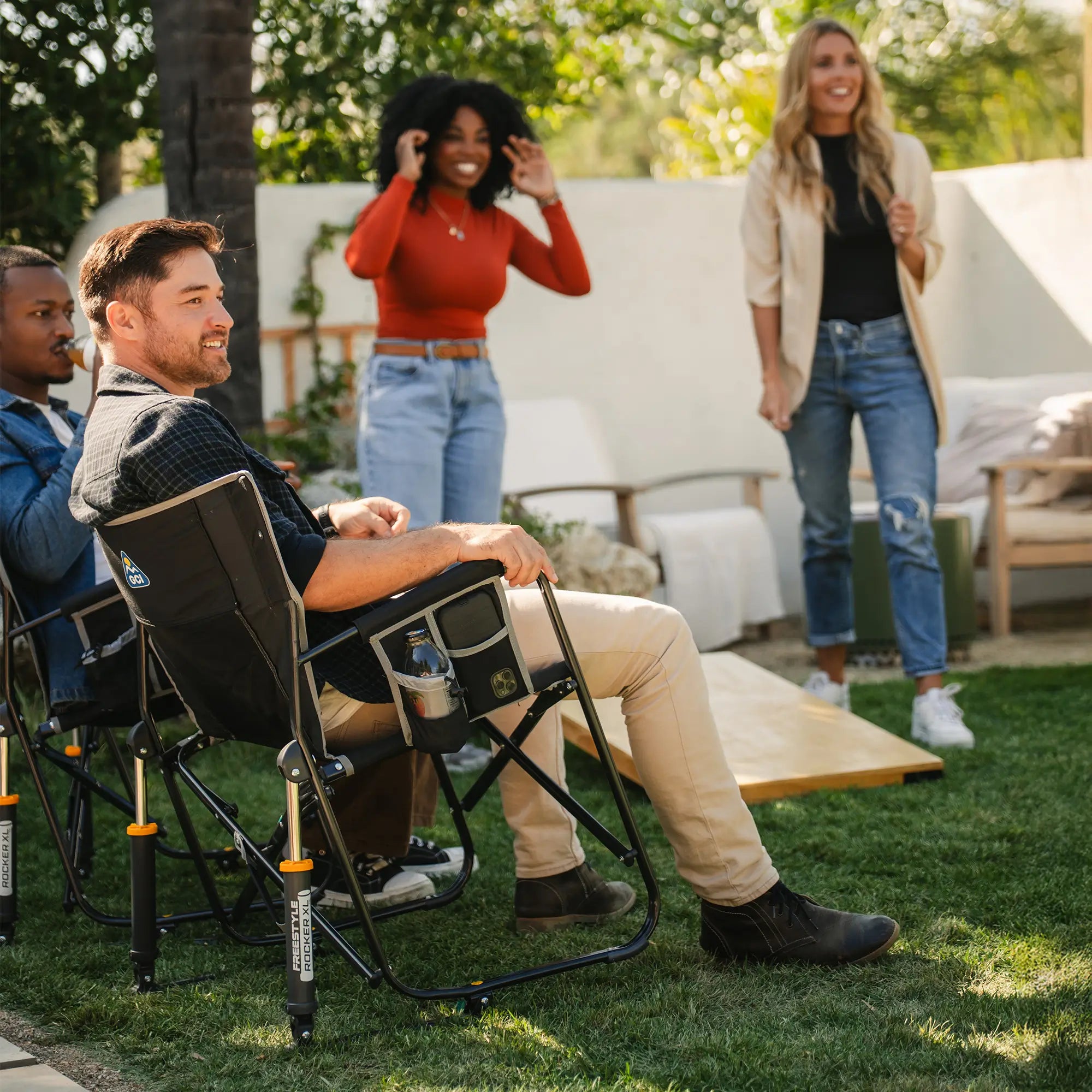 Two people sitting in a black freestyle rocker xl while watching two others play corn hole in front of them. 