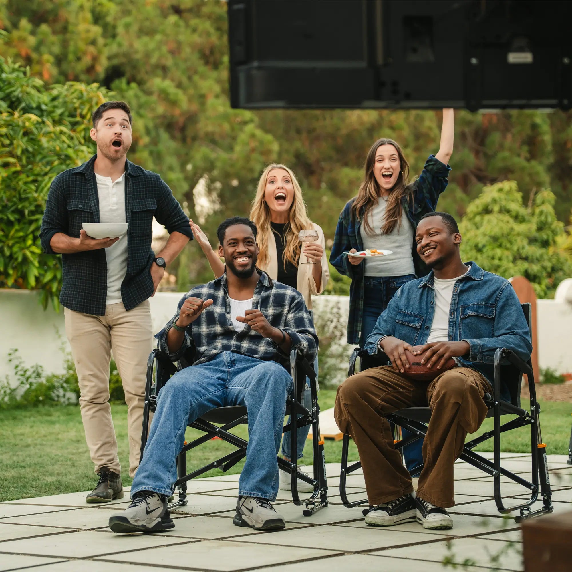 Two men sitting in a black freestyle rocker xl while three people stand behind them cheering at the television. 