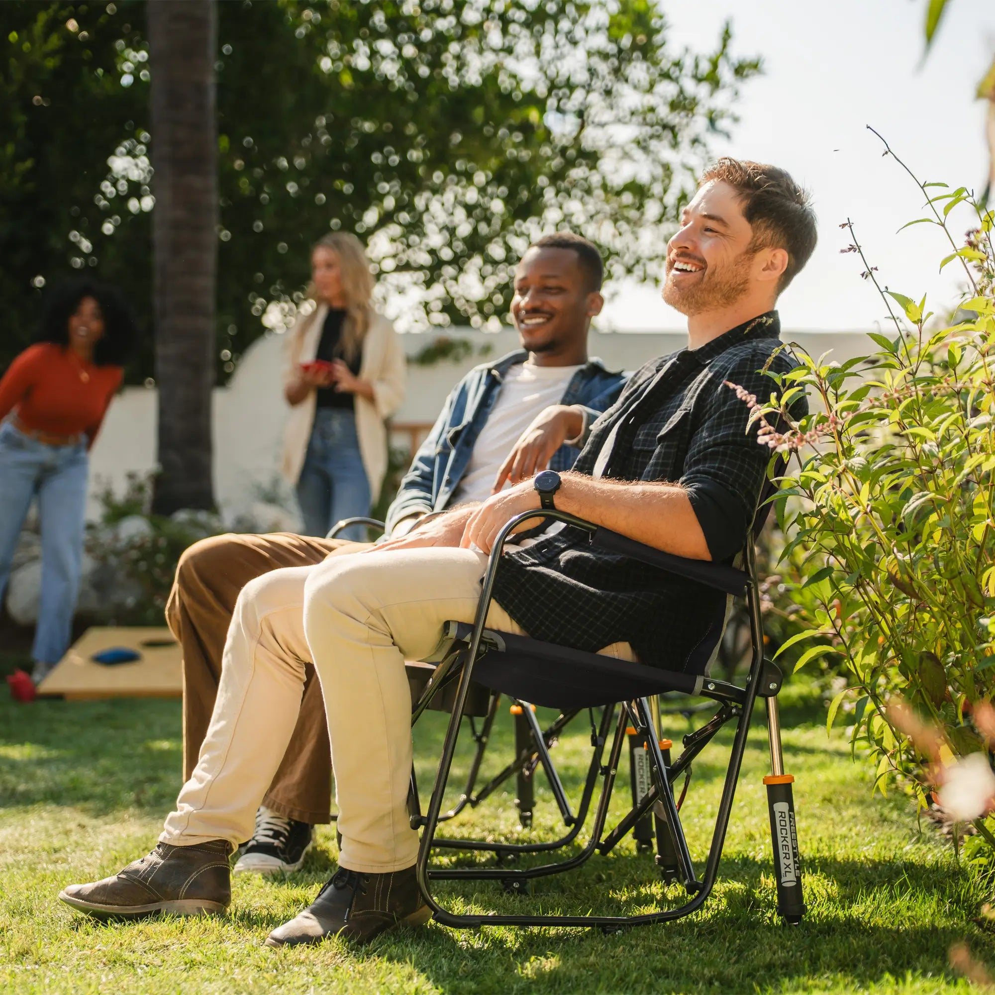 Two men watching people play cornhole while sitting in their freestyle rocker with side table chairs. 