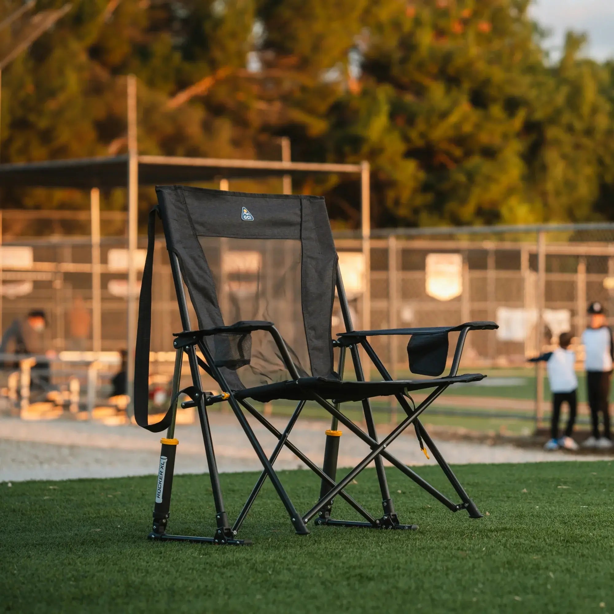 A Comfort Pro Rocker XL chair sits on a grassy field near a baseball game, bathed in warm evening light.