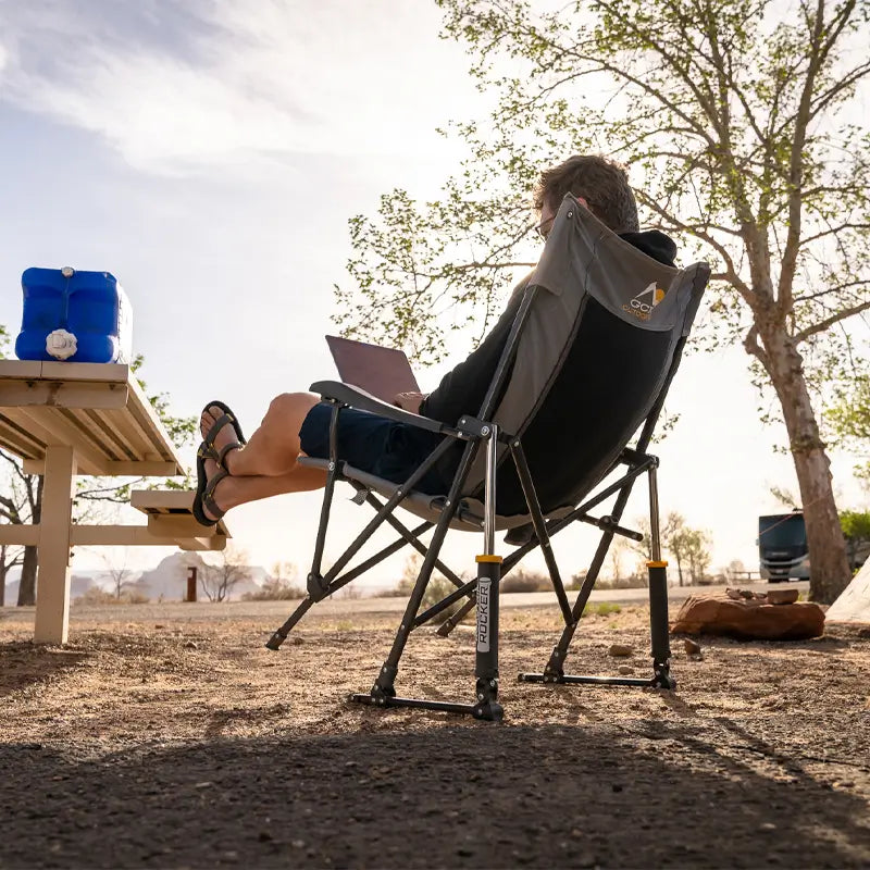 Man relaxes in a Roadtrip Rocker chair, working on a laptop at a picnic area.