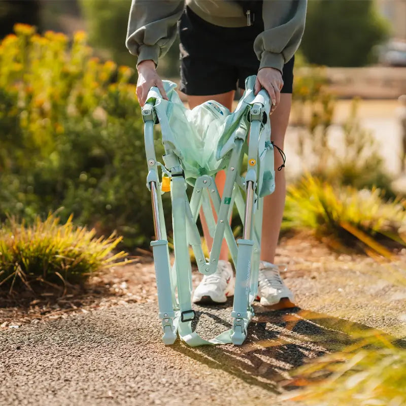 Person unfolding a pastel green Stowaway Rocker outdoors on a path surrounded by plants.