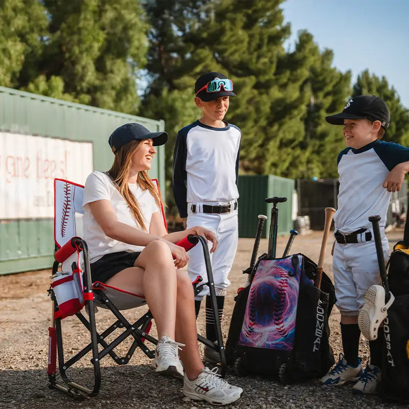Woman sitting on a chair with two young boys in baseball uniforms at a sports field.
