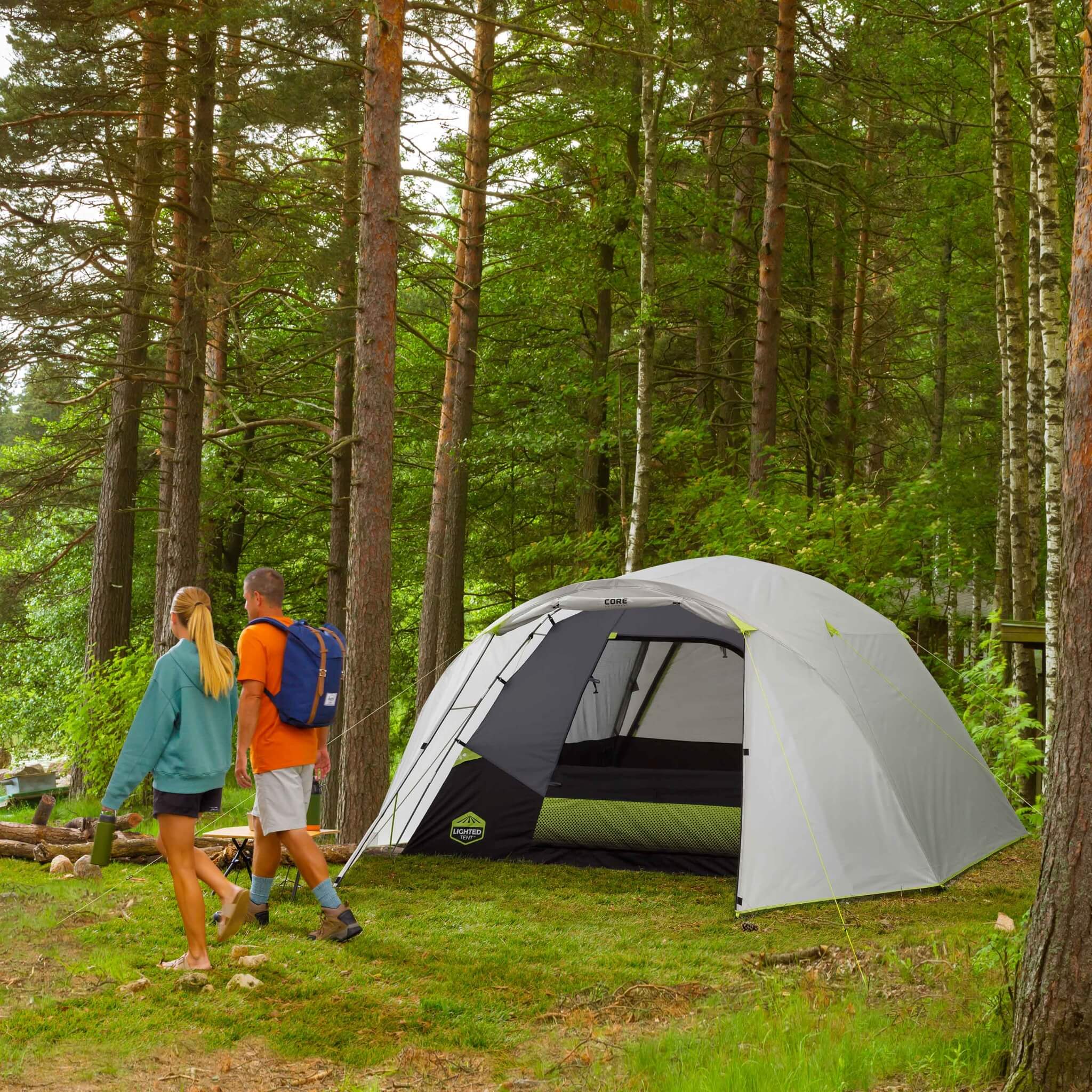 CORE 6 person lighted dome tent with full rainfly set up in a forest clearing, campers preparing for an outdoor adventure.